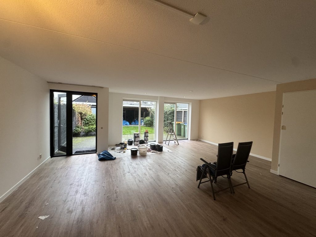 Lege woonkamer met lichte wanden, laminaatvloer en drie glazen deuren naar de tuin, klaar voor de behanger