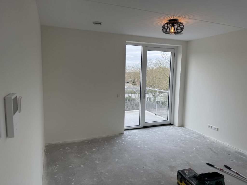 Opgeleverde nieuwbouwkamer met witte wanden, betonvloer en openslaande deuren naar een balkon
