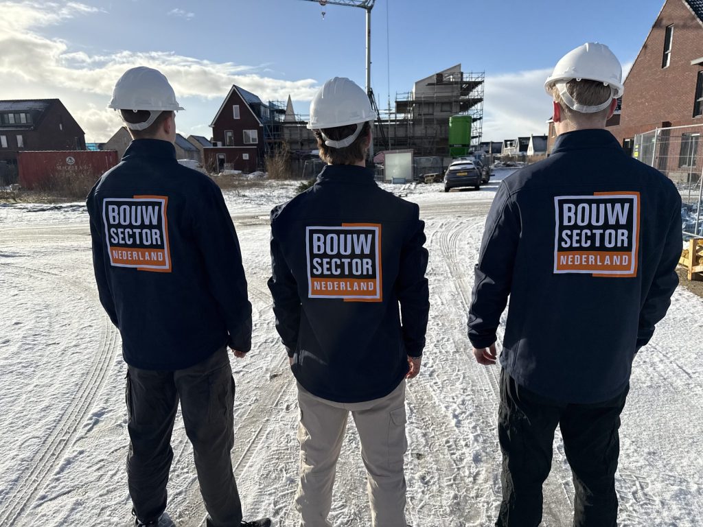 Drie vakmensen van Bouwsector Nederland met witte helm kijken uit over een besneeuwde nieuwbouwwijk in aanbouw
